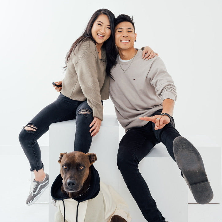 Two people sitting on a white surface with a dog in front of them, all smiling.